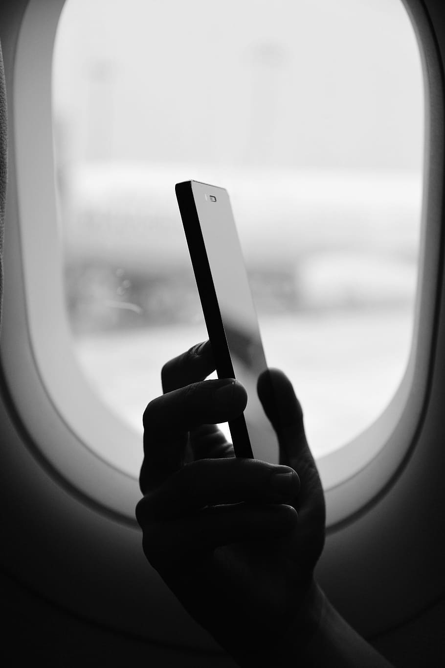 grayscale photography of person holding smartphone beside window, person sitting near airliner window while holding smartphone