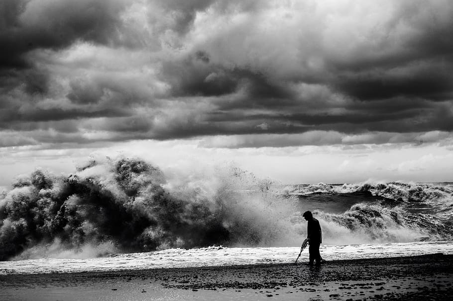 grayscale photo of man walking on seashore, silhouette of man on shore with huge waves and cloudy sky