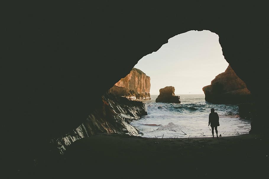 silhouette of person standing under cave near seashore during daytime HD wallpaper
