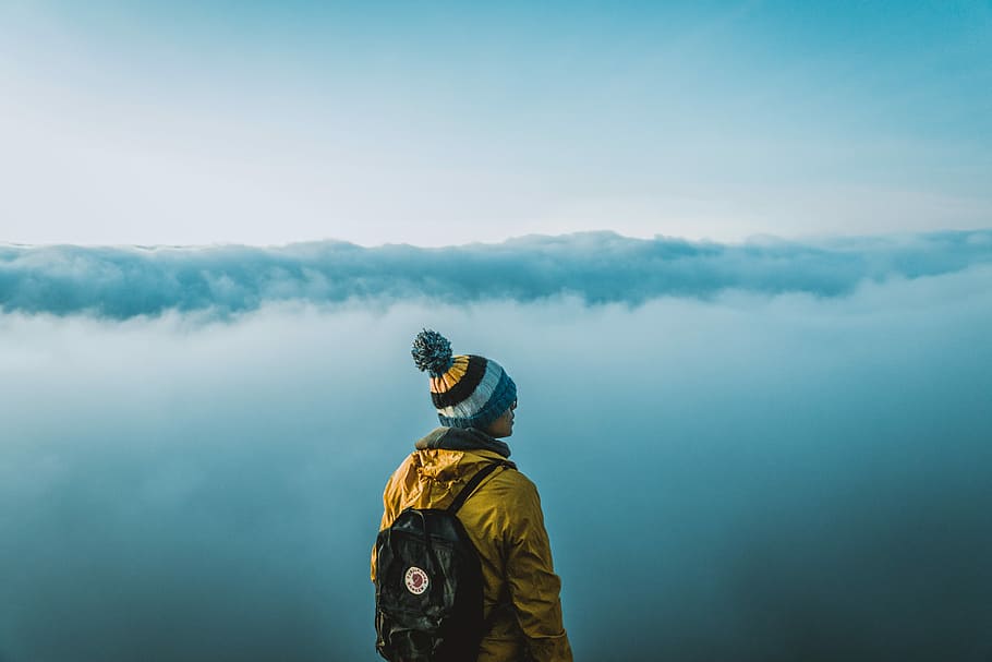 man standing on top of the mountain, person wearing yellow jacket and bubble knit cap standing near white clouds at daytime, HD wallpaper