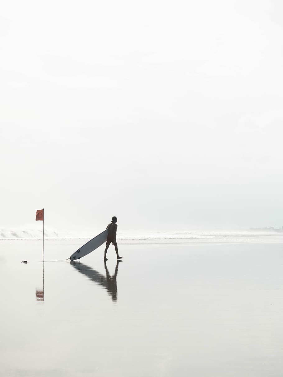 man walking while dragging surfboard at the beach near flag during day, man holding long surfboard walking on shoreline HD wallpaper