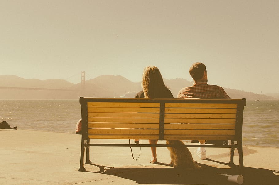 man and woman sitting on bench beside body of water, woman and man seating on brown wooden bench during daytime