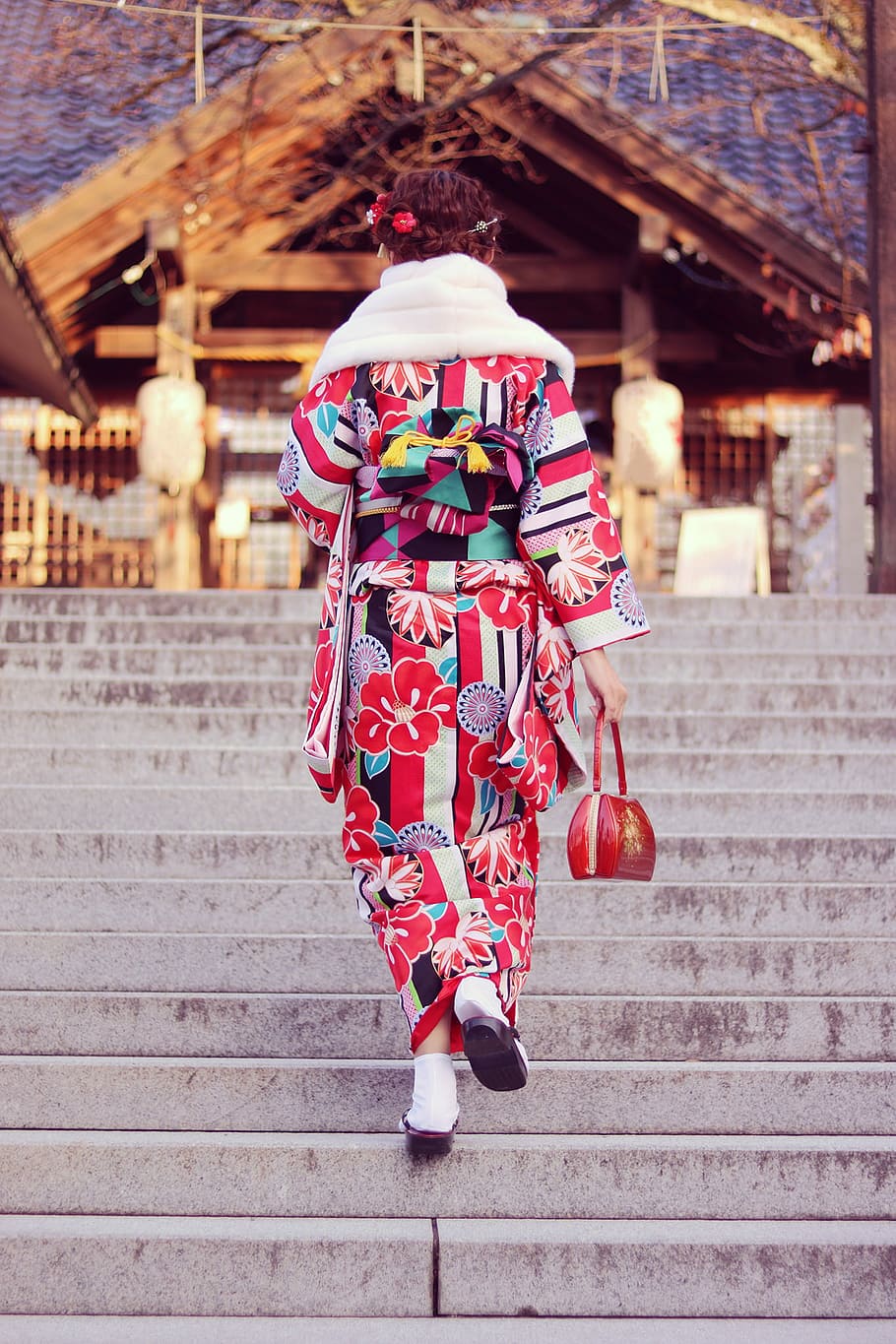 woman in red, white, and black floral long-sleeve traditional dress on stairs facing the brown wooden house