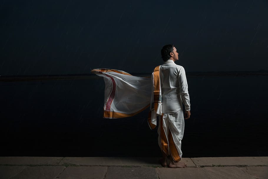 man in white and brown traditional dress standing beside black wall, man standing in front of body of water facing sideways