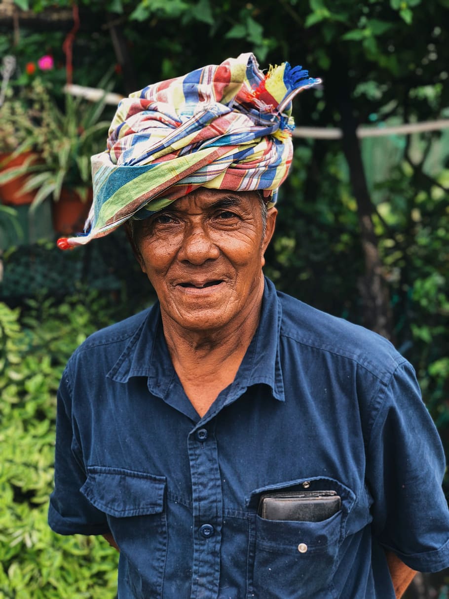 man wearing blue collared shirt standing during daytime photography, man wearing blue button-up collared t-shirt and multicolored headdress standing near green leaf plants during daytime HD wallpaper